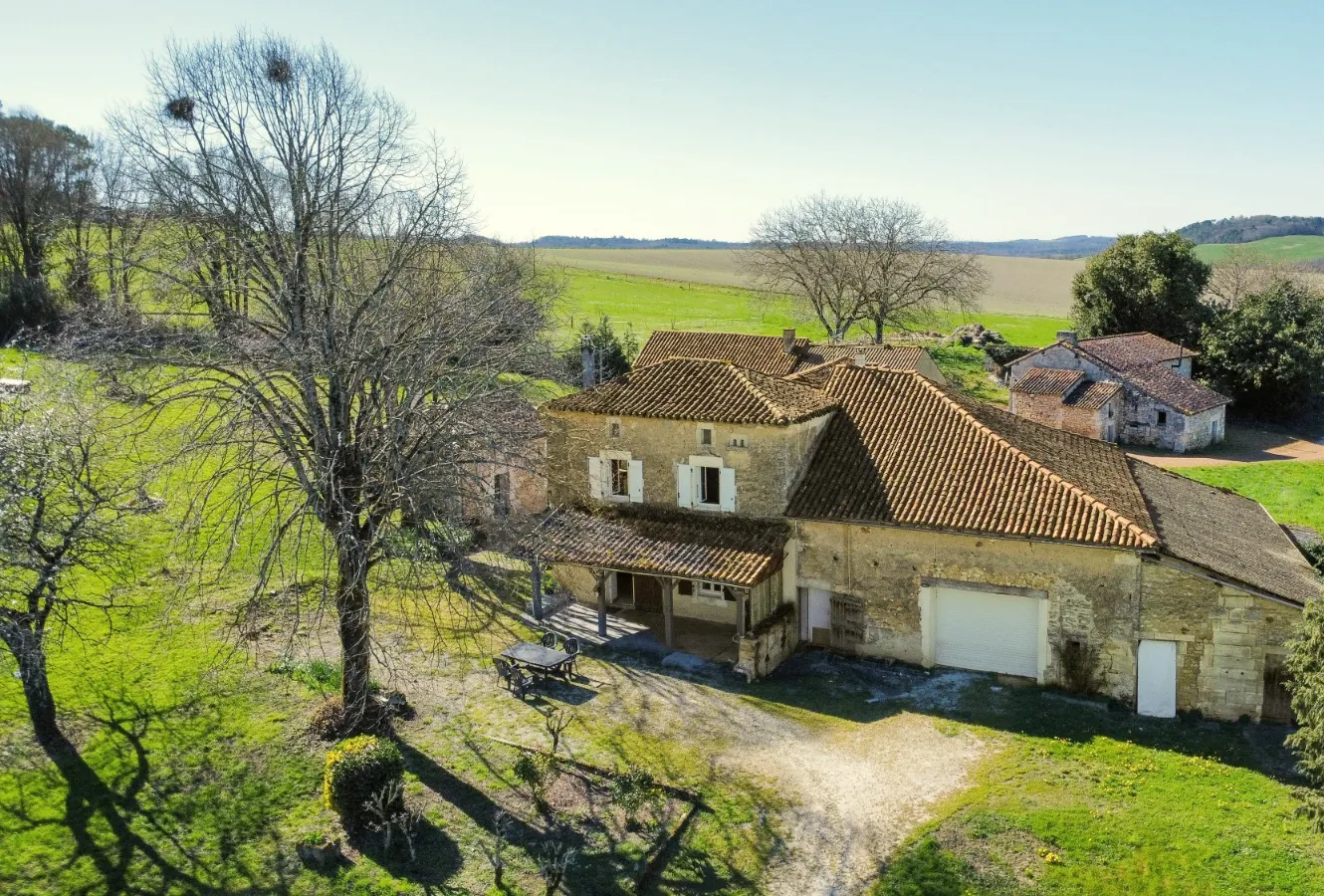 Le gîte vu du ciel au printemps : la ferme en pierre, sa terrasse, le grand arbre et le terrain